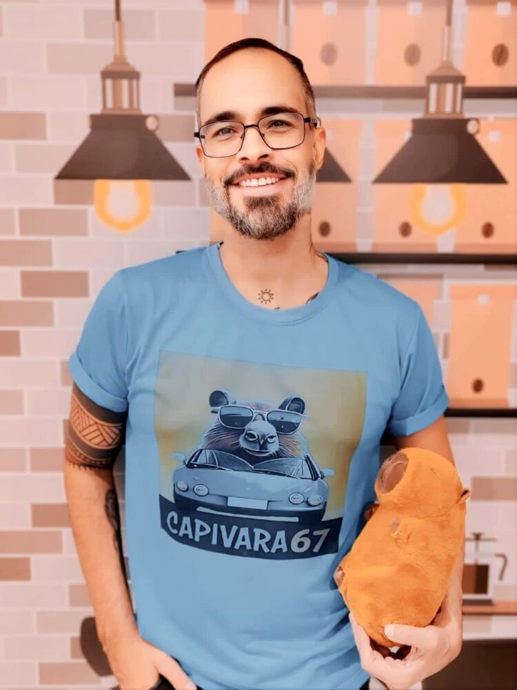 Homem sorridente com óculos e camiseta azul estampada com desenho de capivara dirigindo um carro, segurando um pelúcia de capivara.