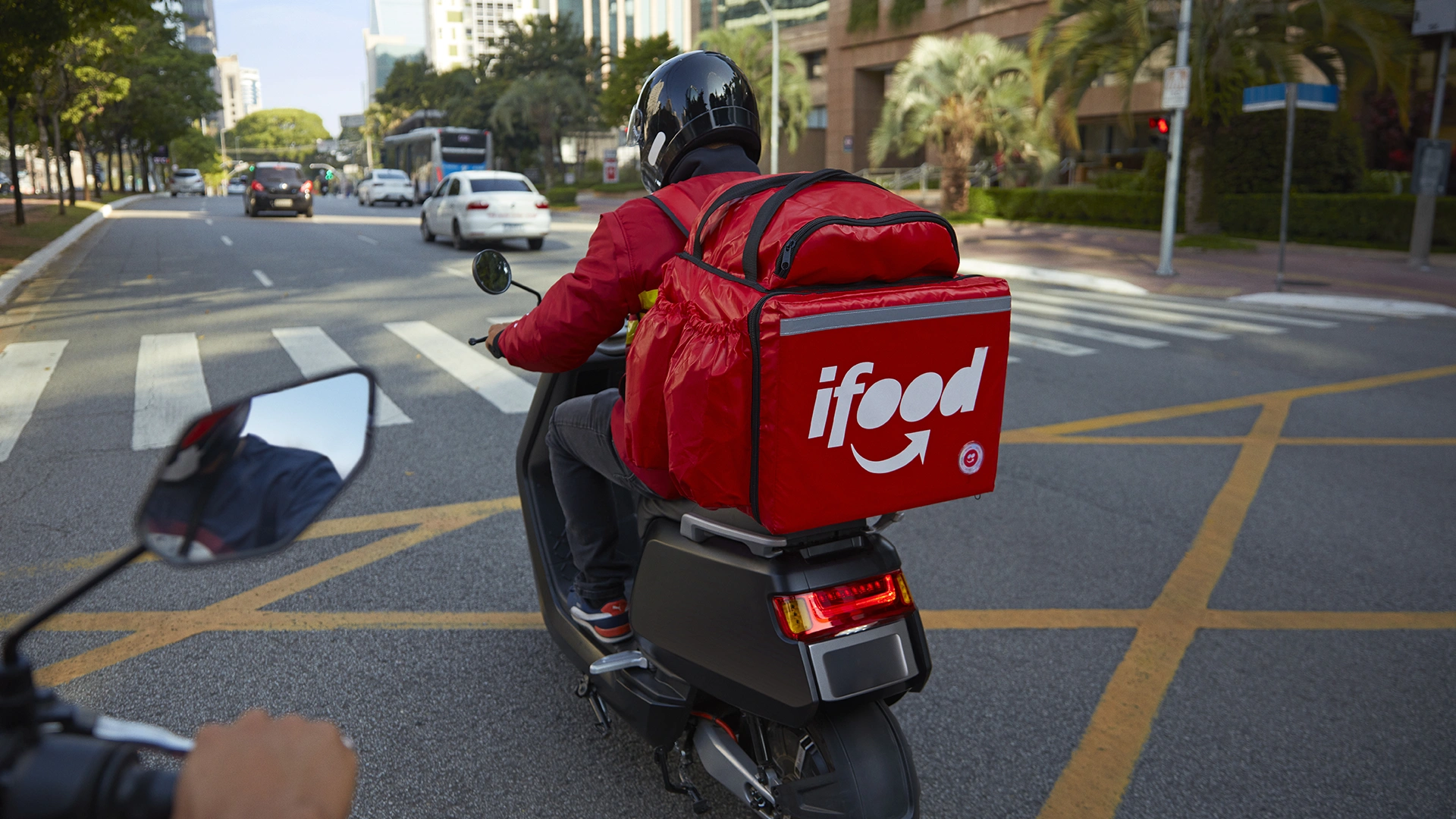 A imagem mostra um entregador do iFood em uma scooter, transitando por uma avenida movimentada de uma grande cidade.