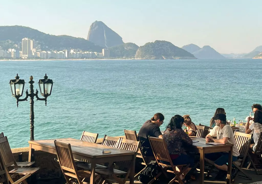 A imagem mostra um grupo de pessoas sentadas em mesas de madeira ao ar livre, com uma vista panorâmica do mar e do Pão de Açúcar ao fundo, no Rio de Janeiro. O cenário inclui o céu claro, montanhas, e o mar azul calmo. À esquerda, um poste de iluminação de estilo antigo aparece em destaque, complementando a atmosfera agradável e descontraída do local. O ambiente sugere um restaurante ou café com mesas externas, onde as pessoas parecem aproveitar uma refeição ou bebida enquanto apreciam a paisagem natural icônica da cidade.