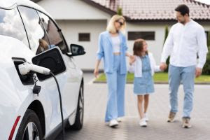"Carro elétrico plug-in sendo carregado em uma estação de recarga com família ao fundo"