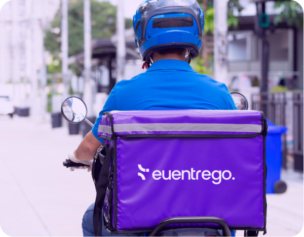 Na imagem, um entregador de moto com bag roxa personalizada da Euentrego realiza uma entrega em área urbana. O serviço de entregas da Euentrego é conhecido por sua eficiência e suporte logístico para negócios. A bag é equipada para garantir o transporte seguro dos produtos.