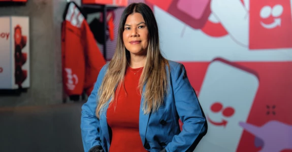 Mulher com cabelos longos, vestindo blazer azul e camiseta vermelha, em um ambiente corporativo com decoração moderna.