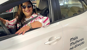 Mulher motorista sorridente ao volante de um carro da 99 com adesivo do programa "Mais Mulheres na Direção".