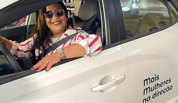 Mulher motorista sorridente ao volante de um carro da 99 com adesivo do programa "Mais Mulheres na Direção".