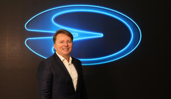 "Flávio Assis, fundador da Lecar, diante de logotipo da empresa em evento corporativo."