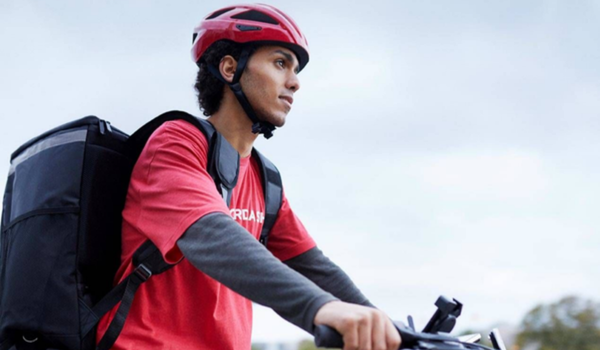 A imagem mostra um entregador usando um capacete vermelho, uma camiseta vermelha e uma mochila de entrega preta nas costas enquanto anda de bicicleta. Ele parece focado e está pedalando em uma área urbana, provavelmente realizando uma entrega. A cena sugere um dia de trabalho típico para entregadores de serviços de entrega rápida.