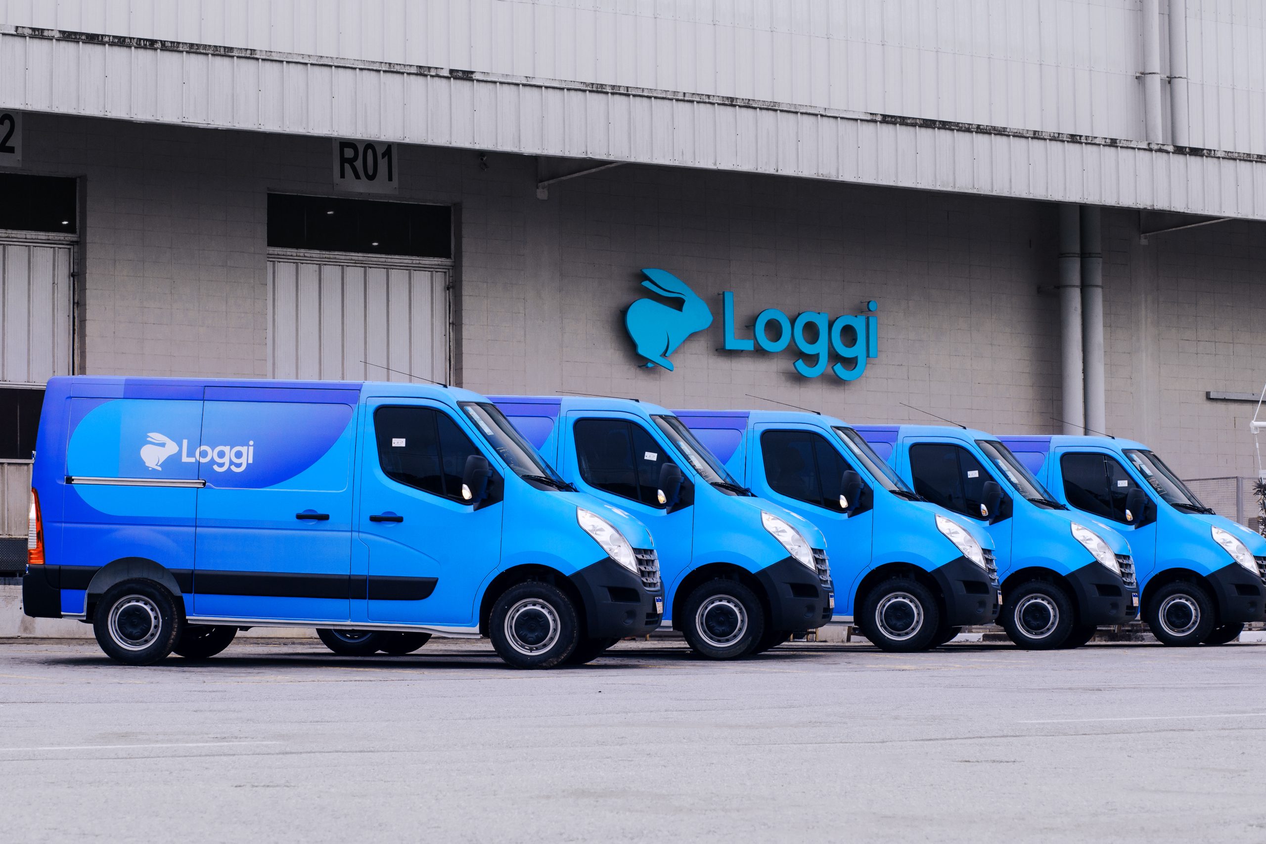 A imagem apresenta uma fileira de vans da Loggi estacionadas em frente a um de seus centros de distribuição. A Loggi, conhecida por seu papel no setor de logística e entregas de última milha, utiliza uma frota moderna para otimizar suas operações de distribuição em grandes centros urbanos. O foco na eficiência logística é evidenciado pela presença dessas vans prontas para atender à crescente demanda de e-commerce e entregas rápidas.