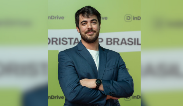 Gabriel West, Ride Hailing Country Lead da inDrive, sorrindo e posando de braços cruzados, vestindo um terno azul-marinho, com um fundo verde contendo a marca inDrive.
