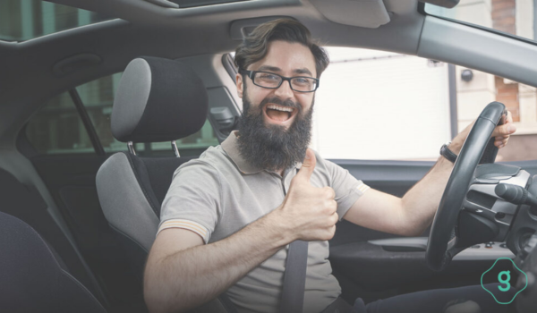 Motorista de aplicativo sorridente com barba e óculos, dirigindo um carro e fazendo sinal de positivo com a mão direita.
