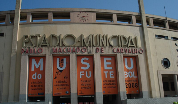 Imagem da entrada do Estádio Municipal Paulo Machado de Carvalho, também conhecido como Estádio do Pacaembu, em São Paulo. Em destaque, há banners laranja com letras brancas formando a palavra "Museu do Futebol", promovendo o museu dedicado à história e cultura do futebol brasileiro. Os textos dos banners mencionam temas como "a ciência, os números, a paixão" e "um espaço para todas as torcidas". A data de inauguração, 2008, também é exibida.