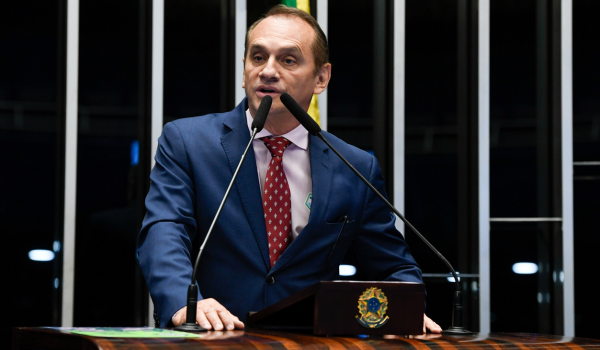 Deputado estadual Wilson Santos discursando em um plenário, vestindo terno azul e gravata vermelha, ao microfone em um púlpito com brasão oficial.