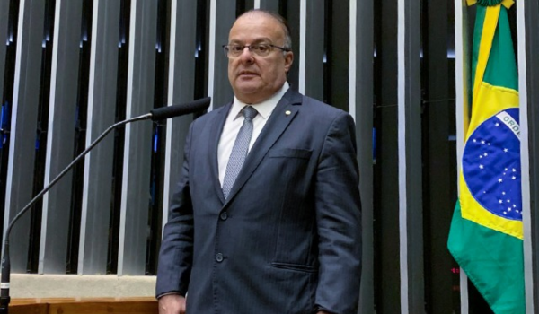 Homem de terno e gravata discursando em ambiente institucional ao lado da bandeira do Brasil.