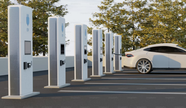 Estação de carregamento para veículos elétricos com seis carregadores alinhados, ao lado de um carro elétrico branco sendo recarregado.