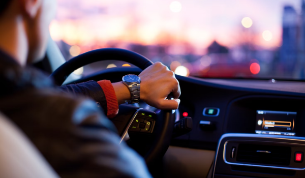 Motorista de aplicativo dirigindo à noite com uma mão no volante, relógio de pulso visível, enquanto o painel do carro está iluminado com o ar-condicionado ligado.