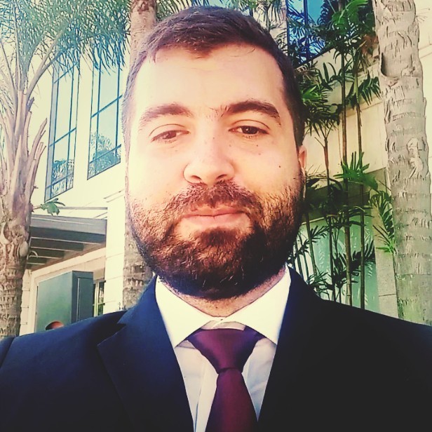 Homem com barba, vestindo terno e gravata, em um ambiente externo com plantas e edifício ao fundo.