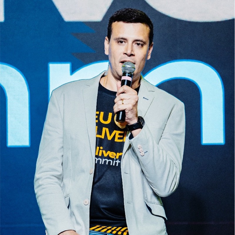 Homem em pé segurando um microfone, usando um blazer claro e uma camiseta com o texto "Delivery Summit". Fundo azul com elementos gráficos.