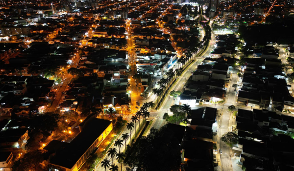 Vista aérea noturna da cidade de Americana, São Paulo, com ruas iluminadas e construções distribuídas ao longo de avenidas cercadas por palmeiras.