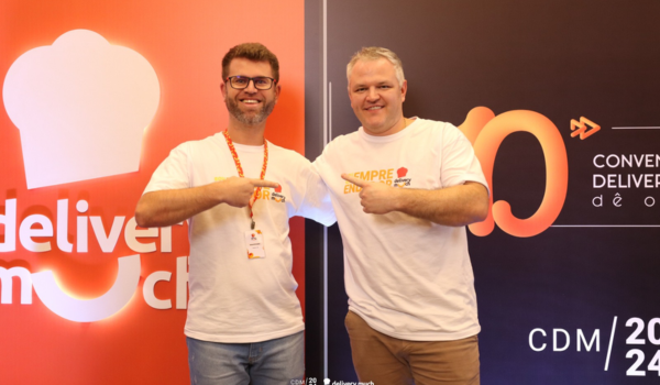 Pedro e Magaiver, ambos usando camisetas brancas do Delivery Much, posam sorridentes em frente a um painel da convenção CDM 2024. Pedro está à esquerda e Magaiver à direita, e ambos apontam um para o outro.