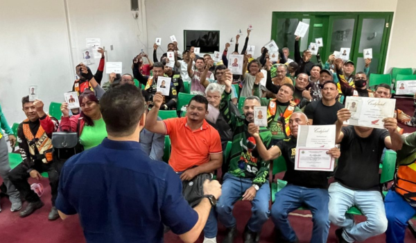 Um grupo de pessoas sentadas em um auditório segura certificados e documentos de identificação, enquanto um homem de costas, usando uma camisa azul, interage com eles.