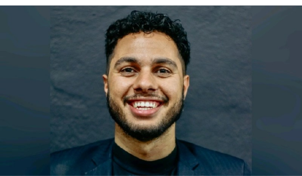Homem sorrindo em um fundo neutro, vestindo blazer preto, representando profissionalismo e carisma.