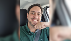 Homem sorrindo dentro de um carro, vestindo camisa verde e com a mão apoiada no queixo.