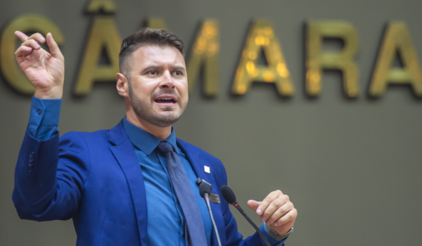 Homem de terno azul discursando em um púlpito com o fundo desfocado onde se lê "CÂMARA".