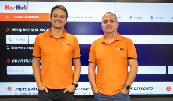 Dois homens posam sorrindo, usando camisetas laranja com o logotipo "KarHub" estampado, em frente a um painel digital com informações sobre peças automotivas.