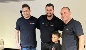 Três homens adultos, todos vestindo camisetas pretas com o logo da Kovi, posam sorrindo ao lado de um menino em um ambiente interno, em frente a uma parede branca.