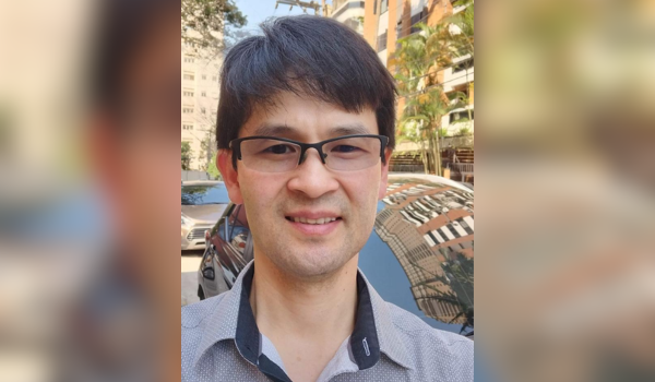 Homem de óculos com expressão sorridente, em ambiente externo com carros e prédios ao fundo.