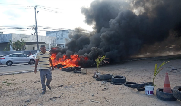 Homem caminha em frente a um protesto com pneus em chamas, gerando uma densa fumaça preta que toma conta do cenário urbano.