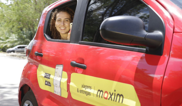 Mulher sorridente com celular na mão, olhando pela janela de um carro vermelho da Maxim, serviço de viagens e entregas.