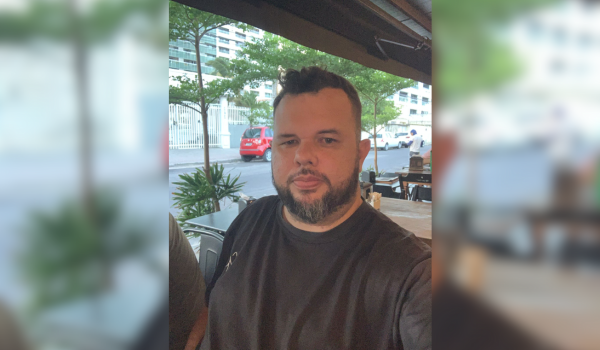 Homem de barba, usando camiseta preta, sentado em um ambiente externo com árvores e prédios ao fundo.