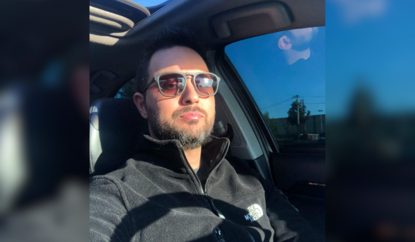 Homem de óculos de sol sentado no banco do motorista, em um carro, vestindo um casaco preto e olhando para a frente.