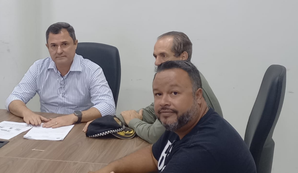 Três homens sentados ao redor de uma mesa durante uma reunião, com documentos e um quepe de policial sobre a mesa.