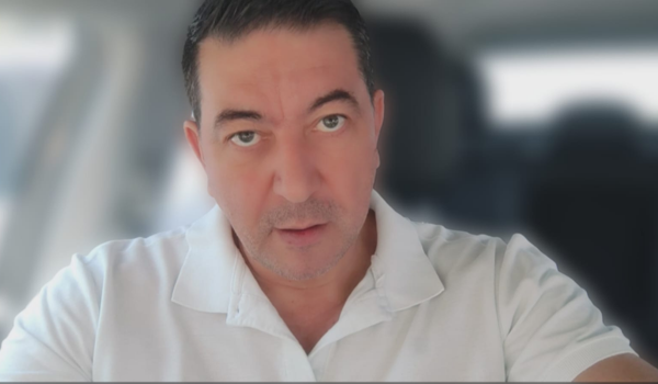 Homem sentado em um carro, vestindo uma camisa polo branca, com expressão séria e olhar direto para a câmera.