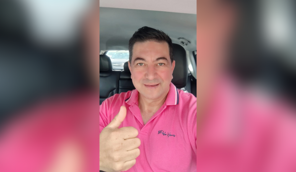 Homem sorridente fazendo sinal de positivo com o polegar dentro de um carro. Ele usa uma camisa polo rosa.