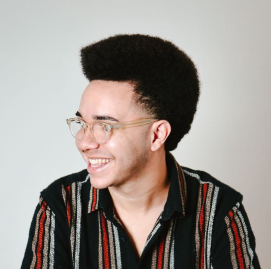 Homem de perfil, com cabelos volumosos, óculos transparentes e camisa listrada, sorrindo e olhando para o lado.