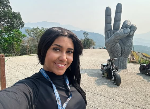 Pessoa sorrindo ao ar livre com uma estátua em formato de mão ao fundo e motocicleta ao lado.