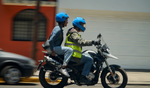 Duas pessoas em uma motocicleta, ambas usando capacetes azuis, sendo uma delas passageira e a outra motorista com colete refletivo.