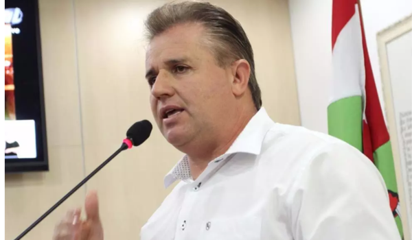 Homem em pé, vestido com camisa branca, fala em microfone em ambiente formal com bandeira ao fundo.