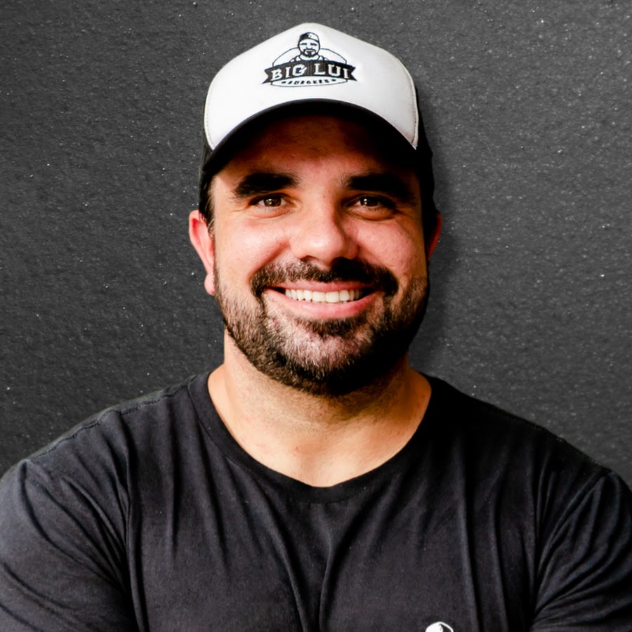 Homem sorrindo, vestindo boné branco com a logomarca "Big Lui Hamburguer", barba curta e camiseta preta, com fundo cinza.