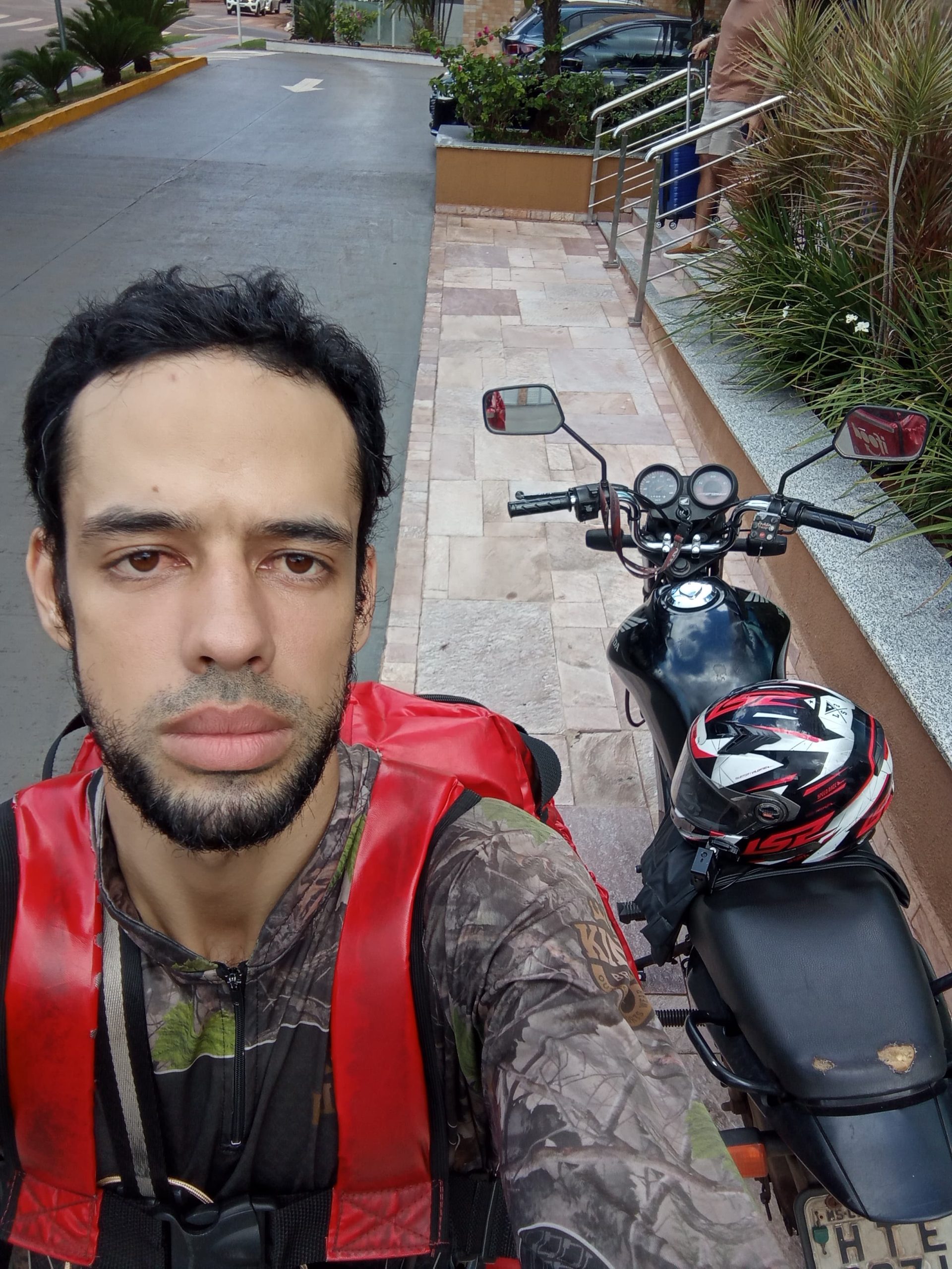 Motociclista parado próximo a uma moto estacionada, com mochila de entregas e capacete no banco.