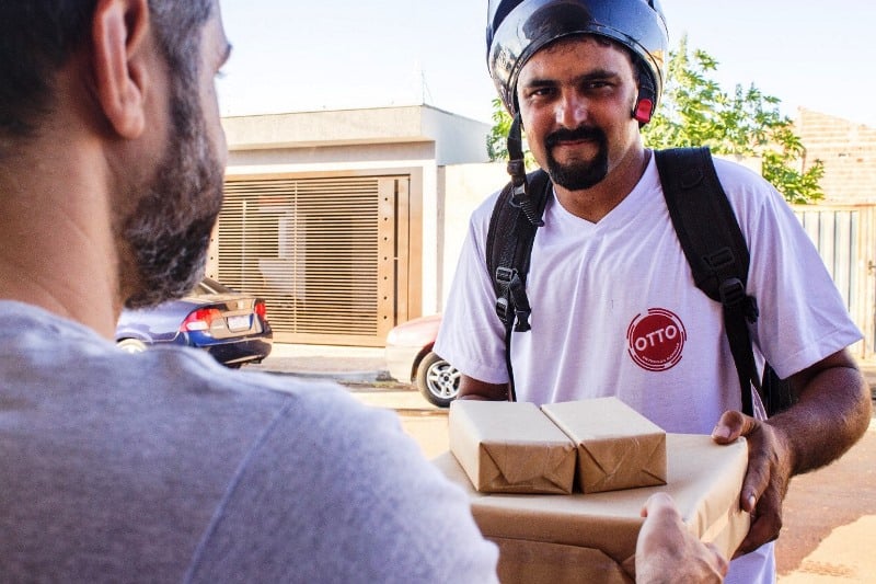 Na imagem, um entregador da Otto Entregas Rápidas, usando capacete e camiseta da empresa, entrega pacotes a um cliente em frente a uma residência. A Otto Entregas Rápidas oferece serviços de entrega para diversos tipos de encomendas.