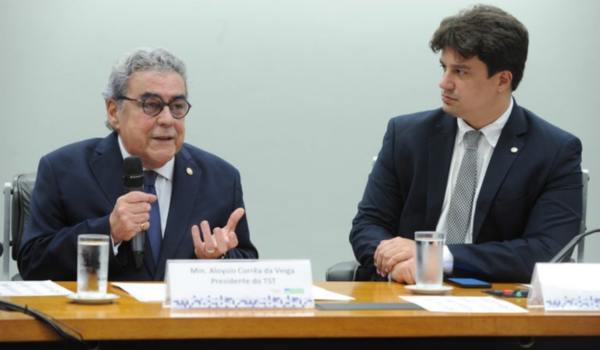 Dois homens de terno e gravata sentados à mesa durante um evento. O homem à esquerda, mais velho, segura um microfone enquanto fala, e o homem à direita o observa atentamente.