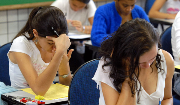 Jovens participando de uma prova em sala de aula, focadas e concentradas, com materiais escolares sobre as mesas.