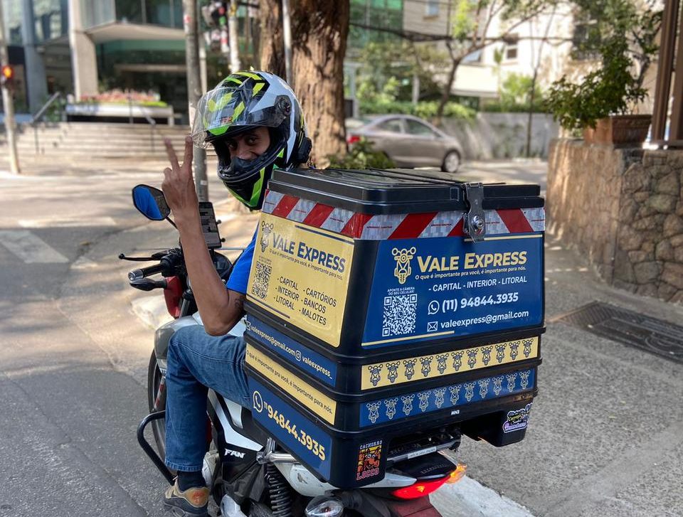 Motociclista com capacete e jaqueta azul conduzindo uma moto com baú da Vale Express em uma rua urbana.