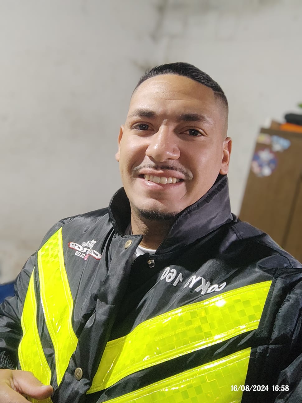 Homem sorrindo, vestindo uma jaqueta preta com detalhes refletivos em amarelo, em um ambiente interno.