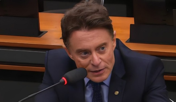 Deputado sentado à mesa, usando terno e gravata, falando ao microfone em uma audiência ou reunião parlamentar.