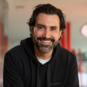 "Homem sorridente com cabelo ondulado e barba, usando uma blusa preta com capuz, posando em um ambiente iluminado com detalhes vermelhos desfocados ao fundo."