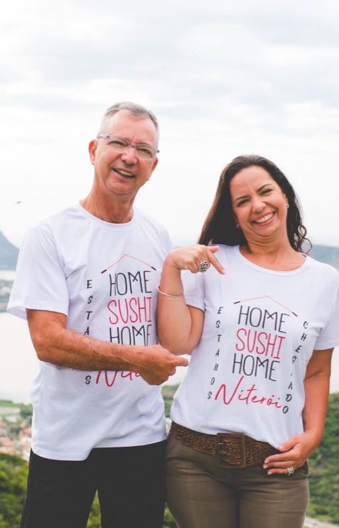 Um homem e uma mulher vestindo camisetas brancas com a frase "Home Sushi Home" e "Niterói", sorrindo e posando para a câmera. Ambos estão ao ar livre, com uma paisagem ao fundo.